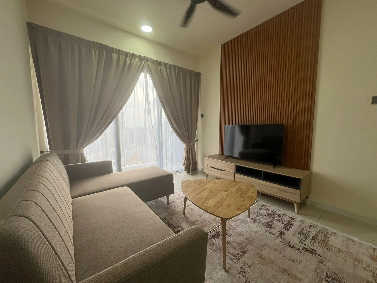 Living features: 3-seater fabric sofa in grey, L-shape sectional sofa, wooden coffee table, TV console with storage, wall-mounted TV, floor-to-ceiling windows, sheer curtains, dark wood accent wall, area rug with geometric pattern, ceiling fan; style: modern; view: city.